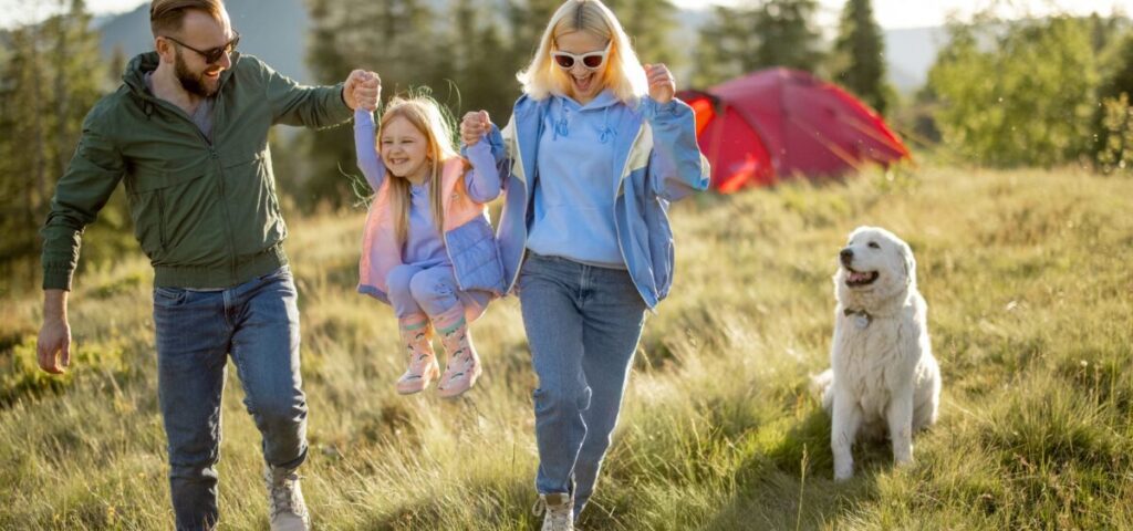 family bonding on a budget - mom and dad swinging girl on a grassy hill with dog looking on